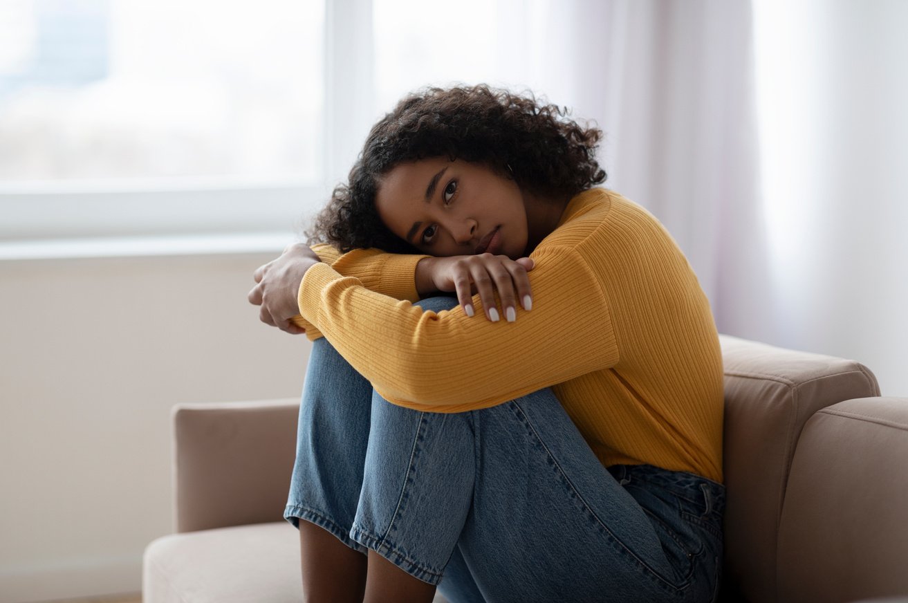 Young depressed black woman hugging her knees on sofa at home, absorbed in negative thoughts, having mental disorder